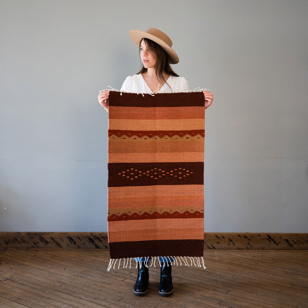 A flat woven wool Oaxacan rug with traditional Oaxacan designed stripes in Maroon, Blush, Terracotta, Rust, Tans. Rug is held up against a grey wall and wood floor by Kelsie in a hat.