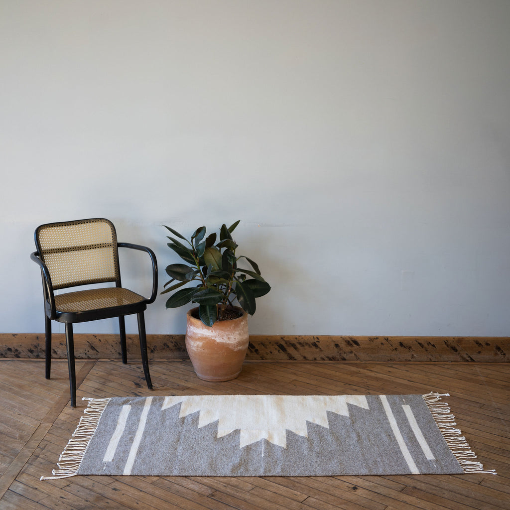A flat woven wool Oaxacan rug with gray background and cream Aztec-inspired pyramid rising off of one side surrounded by one rattan chair and a potted plant. All in front of a gray wall. Wood floors.