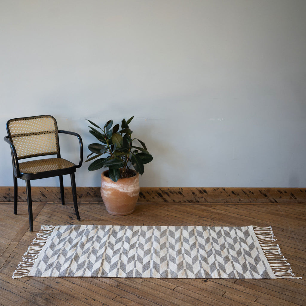 A flat woven cream and light gray Oaxacan rug with a geometric alternating chevron design surrounded by one rattan chair and a potted plant. All in front of a gray wall. Wood floors.