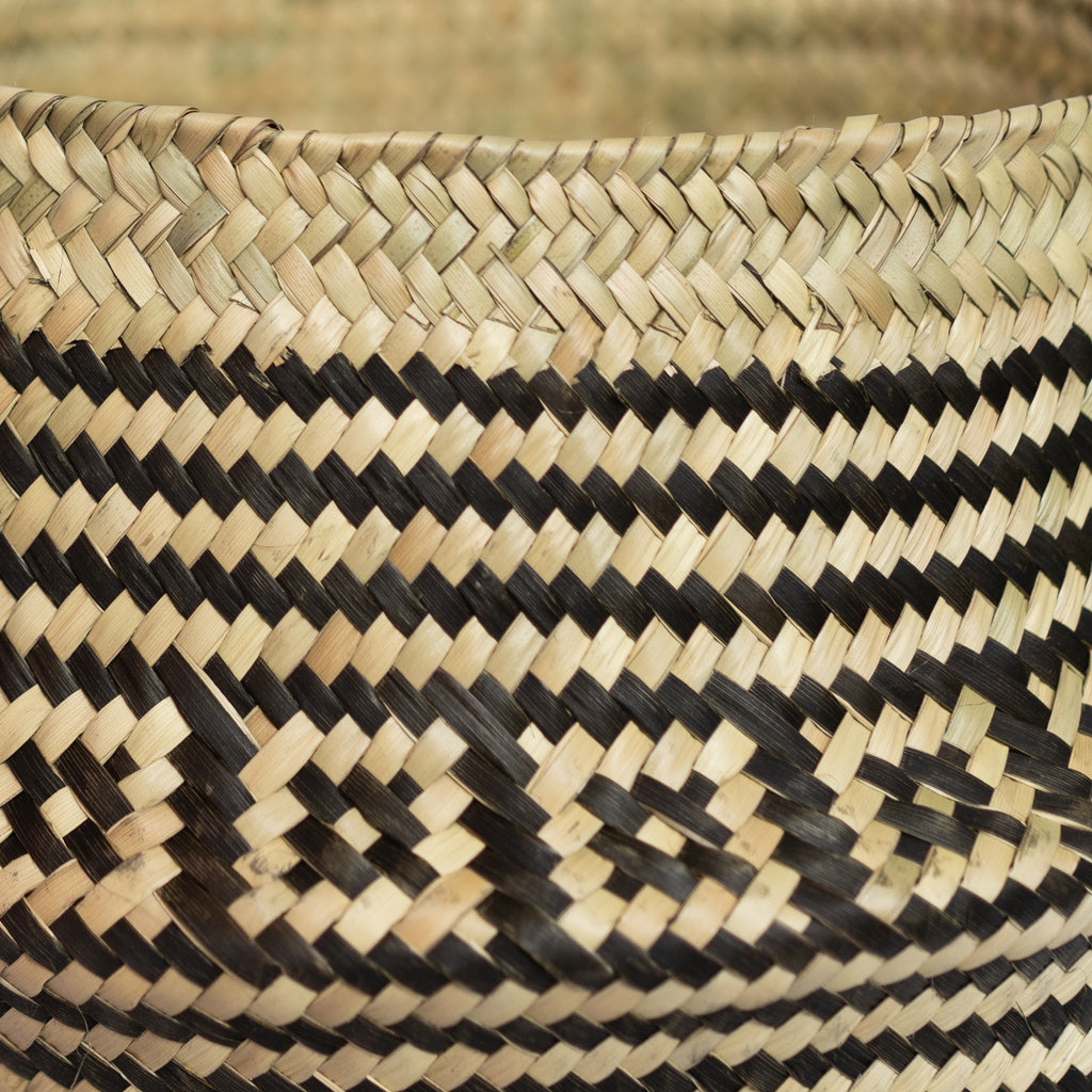 Close up of handwoven palm fiber belly basket in traditional Oaxacan designs. Tan and black stripes alternate with rows of contrasting layered squares. Gray background. 