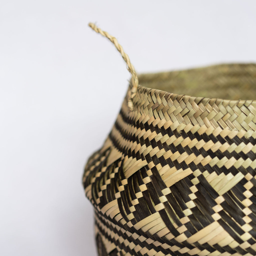 Close up of handwoven palm fiber belly basket with handles in traditional Oaxacan designs. Tan and black horizontal stripes alternate with rows of graphic coffee beans. Horizontal stripes run along both top and bottom edge. Gray background.