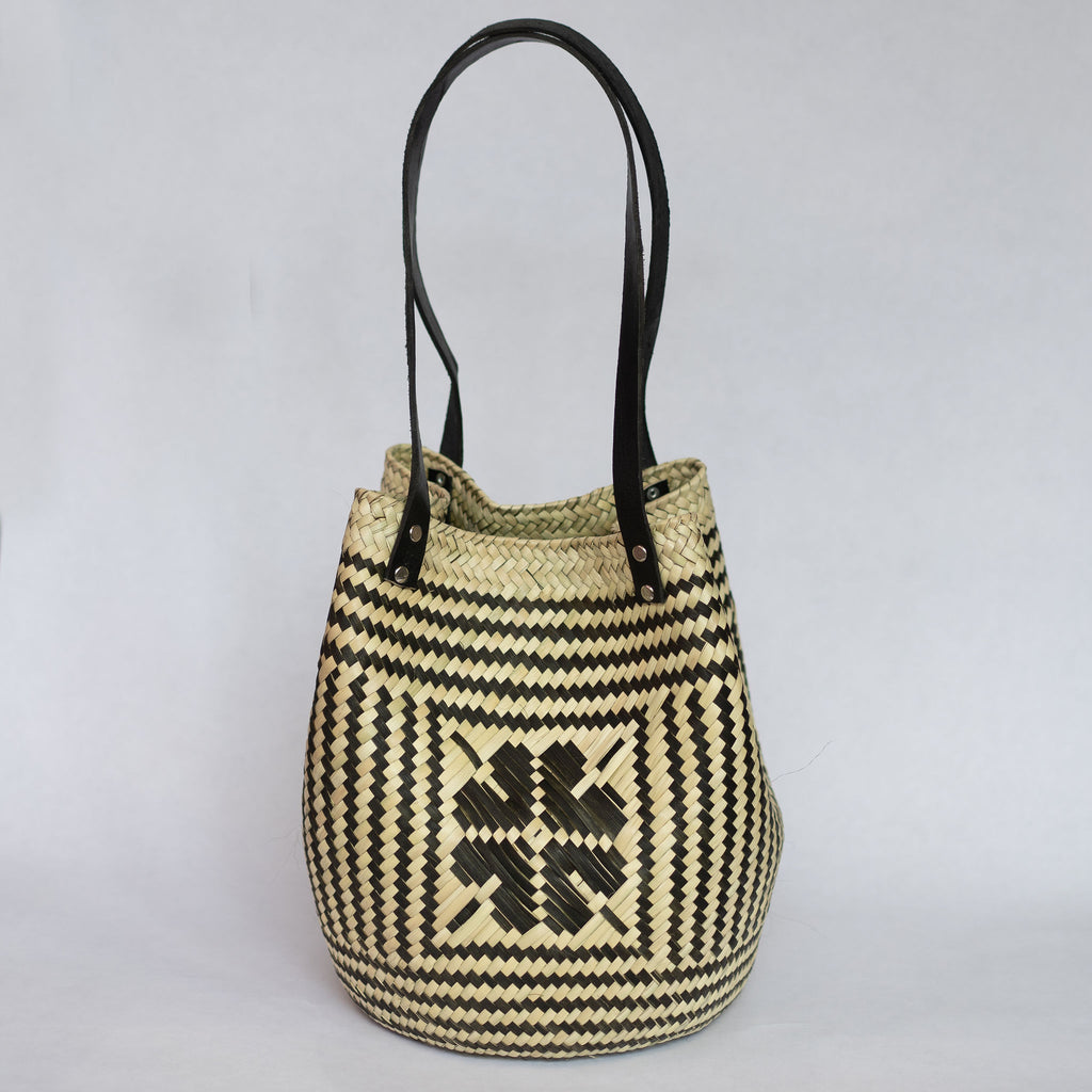Handwoven palm fiber tote bag with black leather handles in traditional Oaxacan designs. Tan and black vertical striped design with flowers on each side. Three black horizontal stripes at top and bottom edge. Gray background.