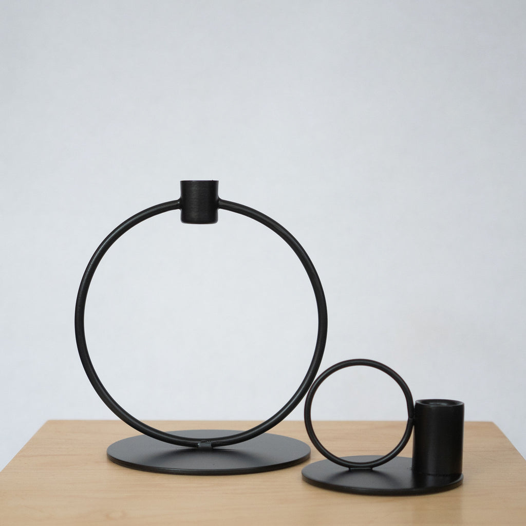 Powder coated iron black taper candle holders. On the left is a large circle with a flat round base and a small cup for the candle at the top. On the right is a small circle set next to a cup for a taper candle all on a small round base. Both sit on a wood platform in front of a light gray background.