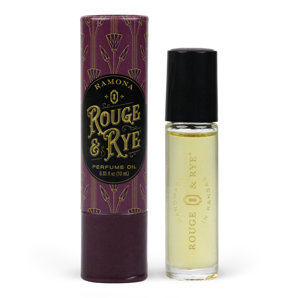 Golden Ramona perfume oil roller in clear glass bottle with black plastic cap next to red/purple Art Deco inspired packaging. White background.