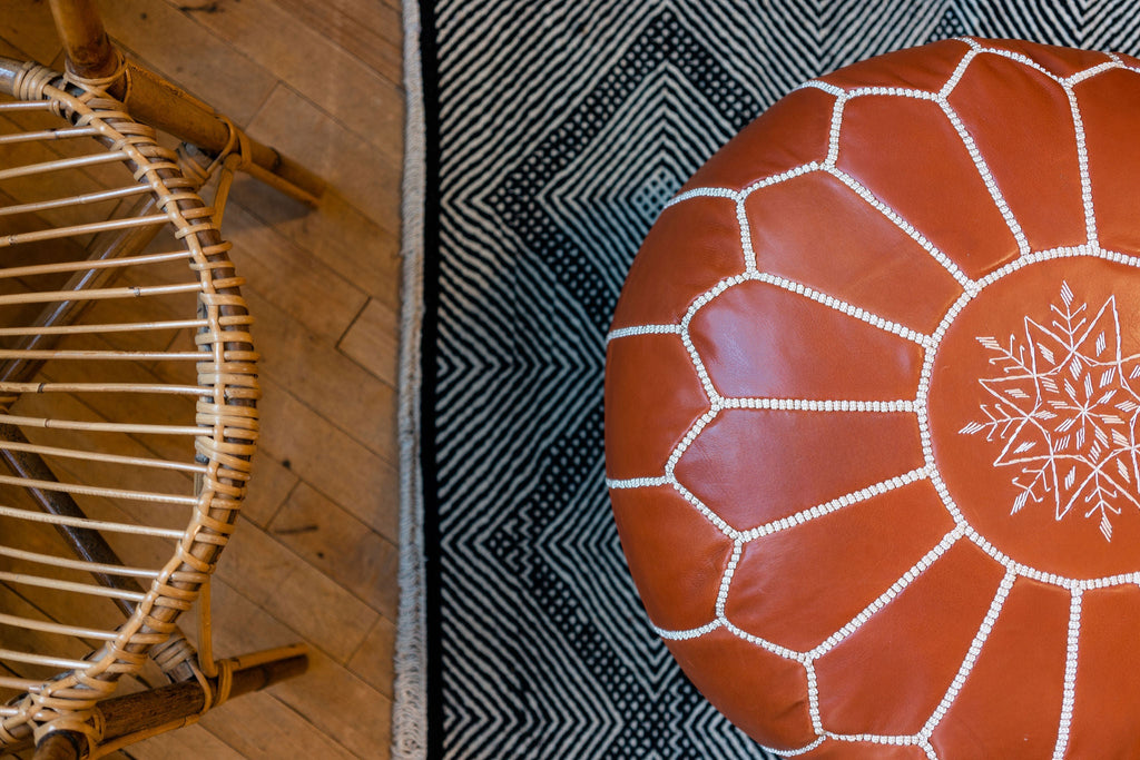 Looking down on the top of a Round Camel Colored Leather Pouf / Ottoman. The embroidery on the top of the pouf is a symmetrical, radial design, with a design reminiscent of a snowflake shape right in the center. Pouf sits on black and white rug next to a rattan chair that peaks in the side of the photo.