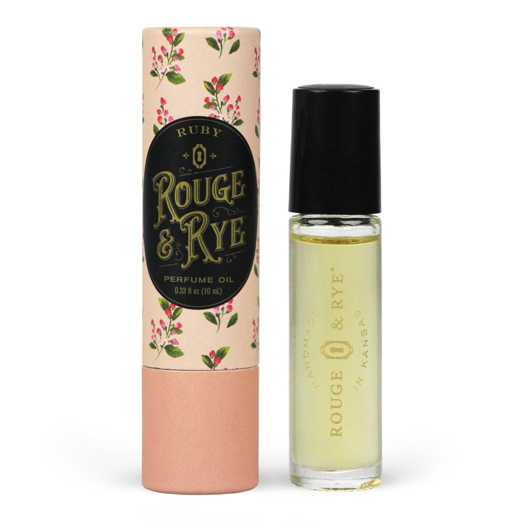 Golden Ruby perfume oil roller in clear glass bottle with black plastic cap next to peach floral packaging. White background.