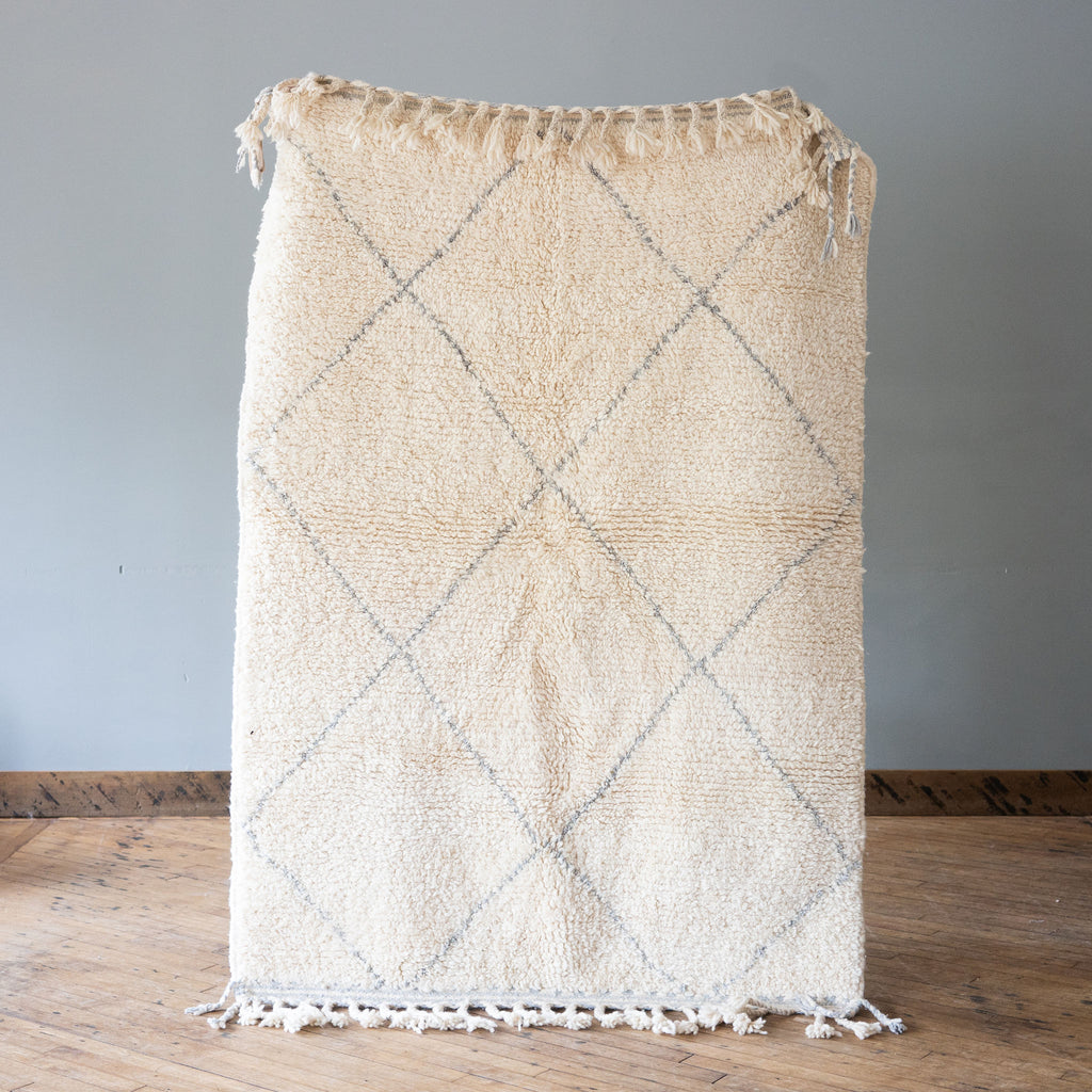 A high pile Moroccan Beni Ourain rug with classic cream and gray diamond design and tasseled edge. Rug is held up against a grey wall and wood floor.