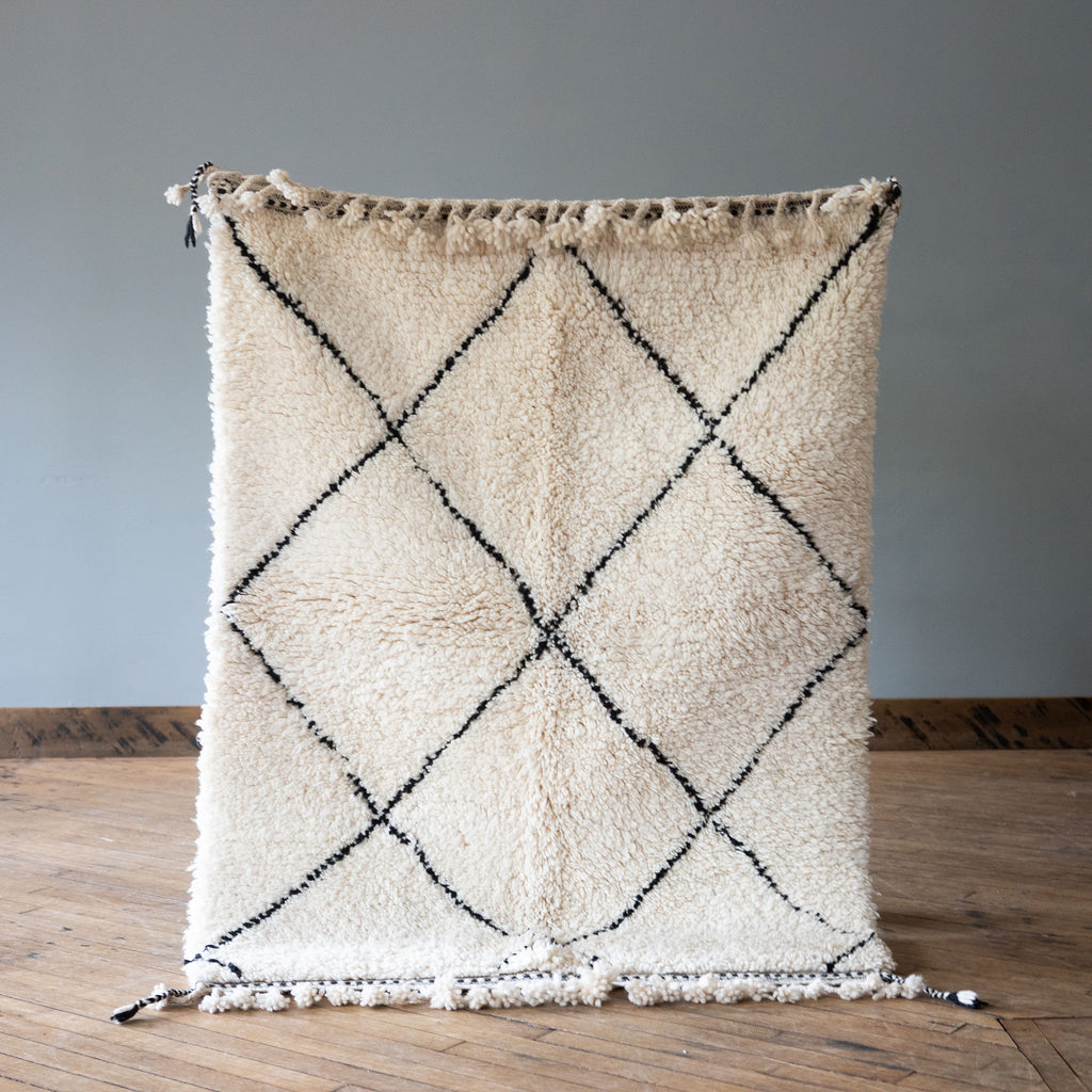 A high pile Moroccan Beni Ourain rug with classic cream and black diamond design and tasseled edge. Rug is held up against a grey wall and wood floor.