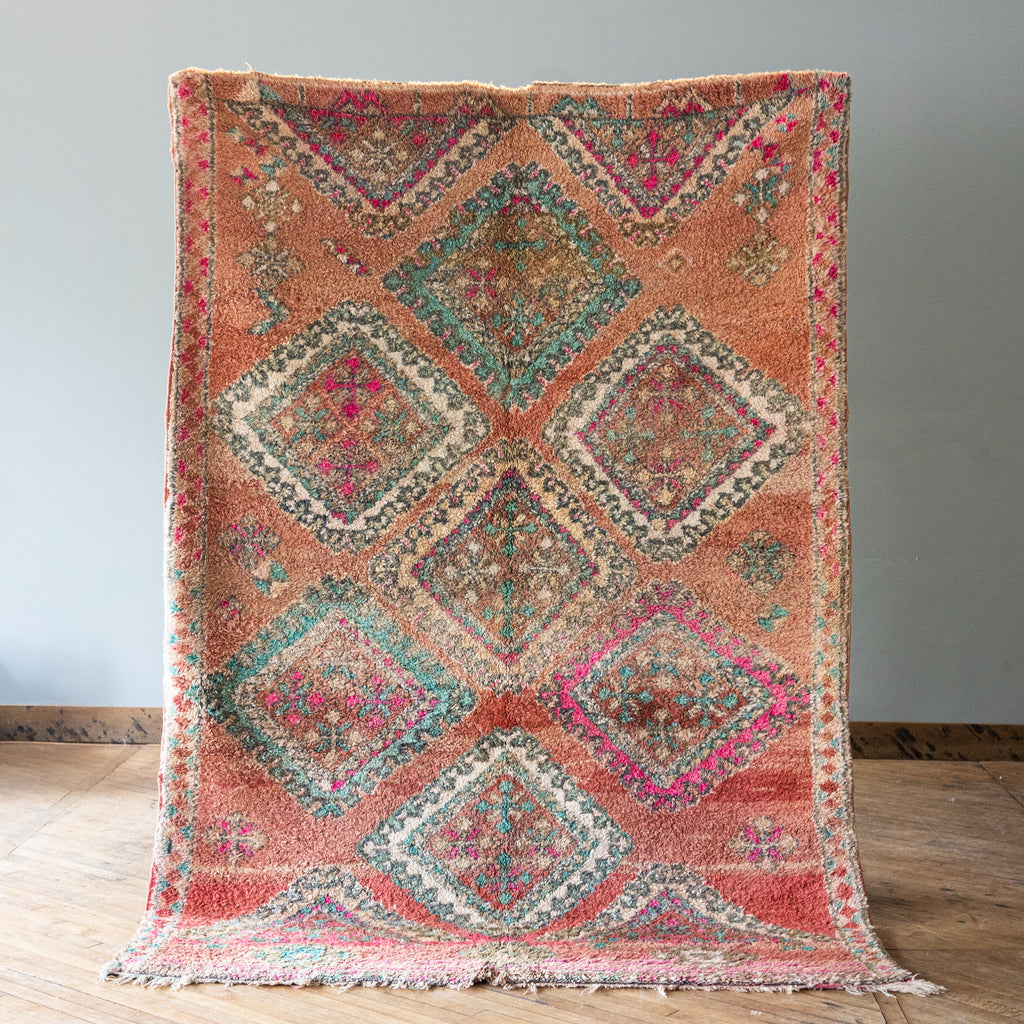 A vintage Moroccan Benimguild rug with a bold diamond pattern in coral, turquoise, cream and pink. Rug is held up against a grey wall and wood floor.