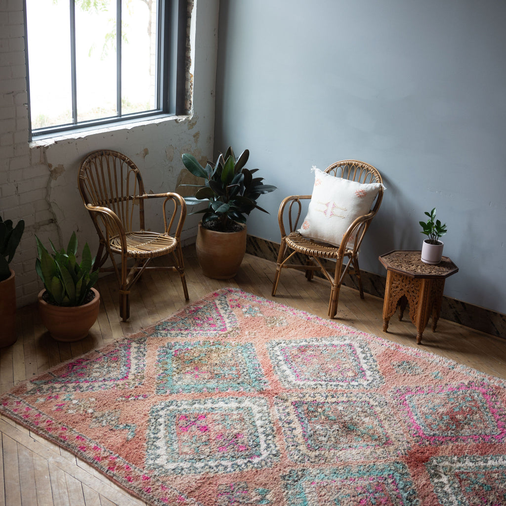 A diagonal view of a vintage Moroccan Benimguild rug with a bold diamond pattern in coral, turquoise, cream and pink surrounded by two rattan chairs, a side table, and a potted plant. Wood floors.