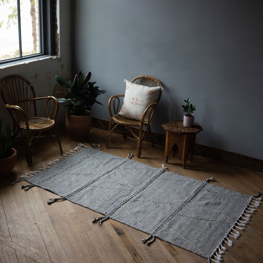 A diagonal view of a flat woven wool Moroccan Zanafi rug with a subtle stripe design in slate gray and white with thicker cords woven through in a large triangle design surrounded by two rattan chairs, a side table, and a potted plant. Wood floors.