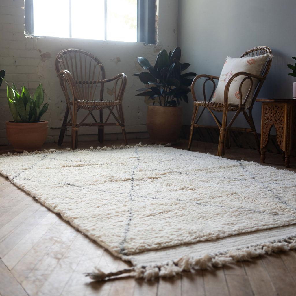 A diagonal view of a high pile Moroccan Beni Ourain rug with classic cream and gray diamond design and tasseled/fringed edge surrounded by two rattan chairs, a side table, and a potted plant. Wood floors.