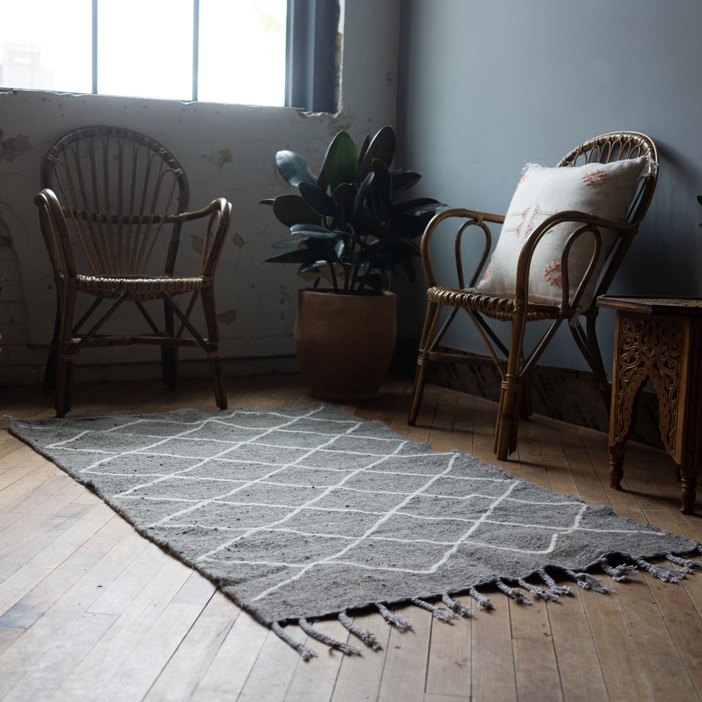 A diagonal view of a flat woven wool Moroccan Beni Ourain rug with gray background and cream diamond designk surrounded by two rattan chairs, a side table, and a potted plant. Wood floors.