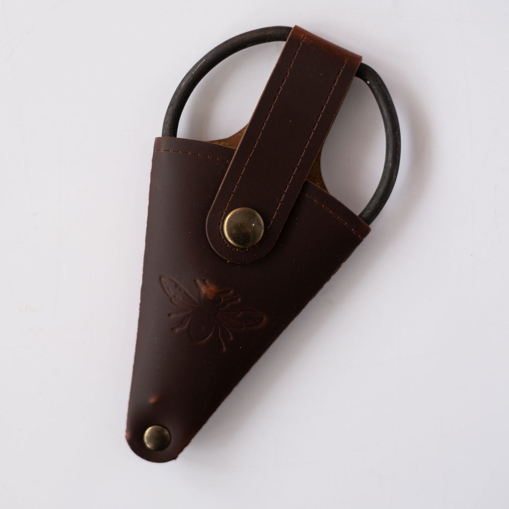 Wide handled loopy scissors inside leather pouch snapped close. Bee engraved on leather pouch. White background.