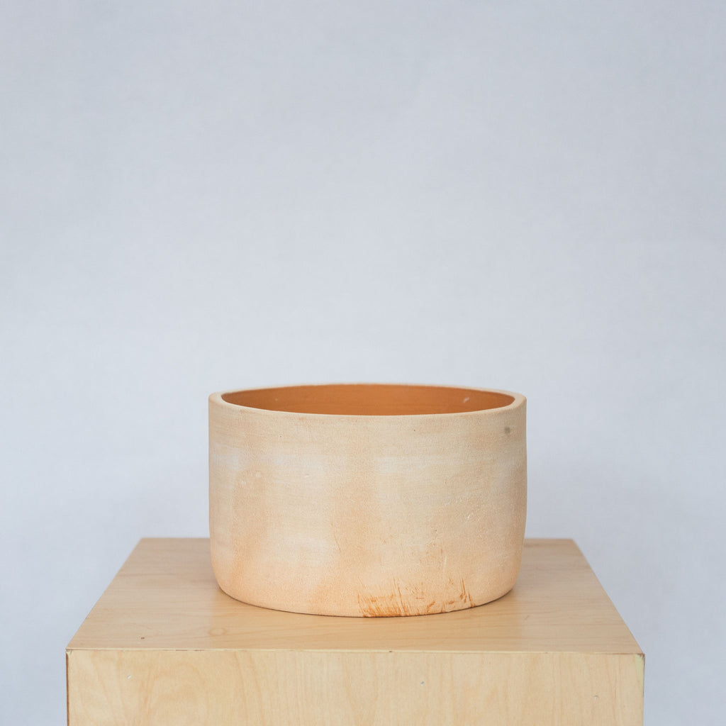 Natural terra cotta toned clay large bowl sits on a wood platform in front of a gray background.