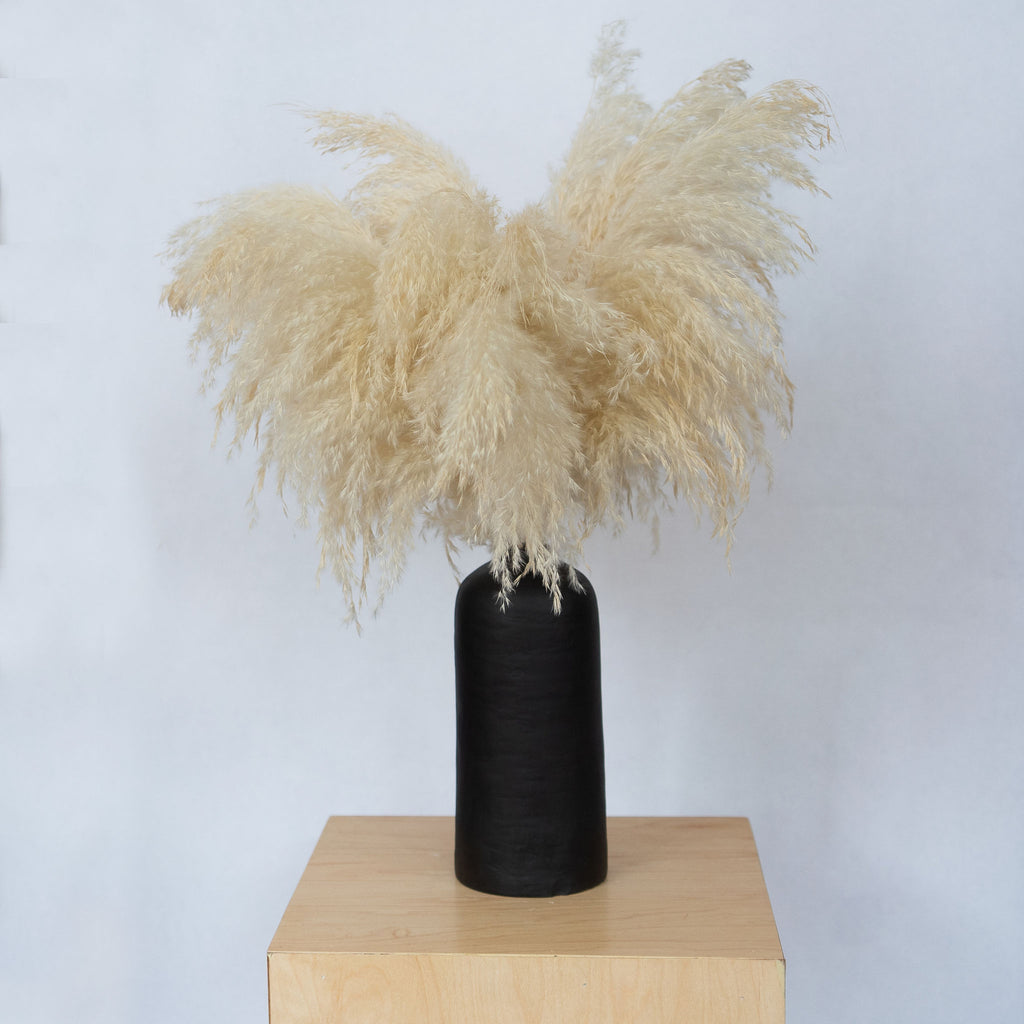 A tall straight sided black ceramic vase holding cream feathers sits on a wood platform in front of a light gray background.