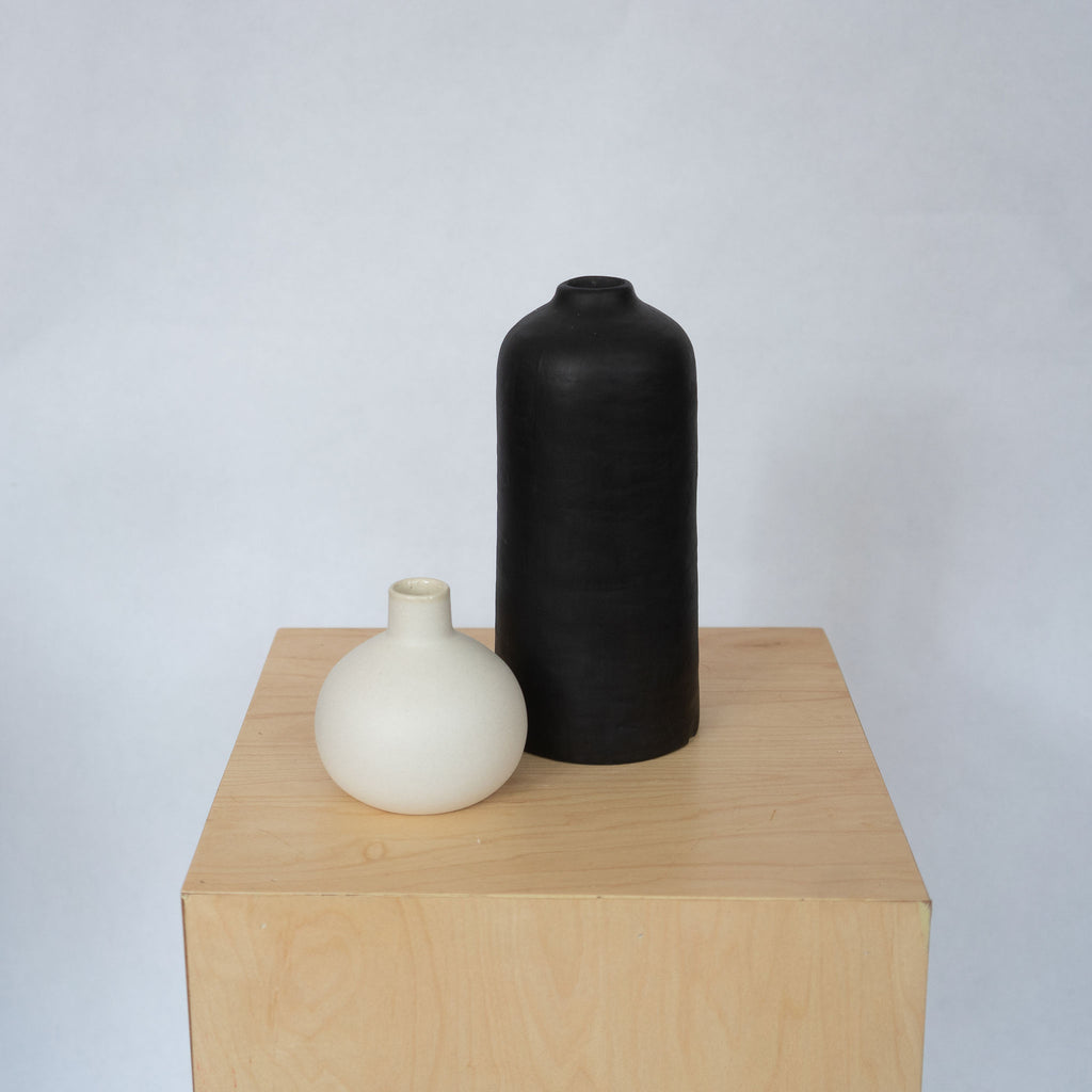 A small round white ceramic vase with skinny neck sits with a taller straight sided black vase on top of a wood platform in front of a light gray background.
