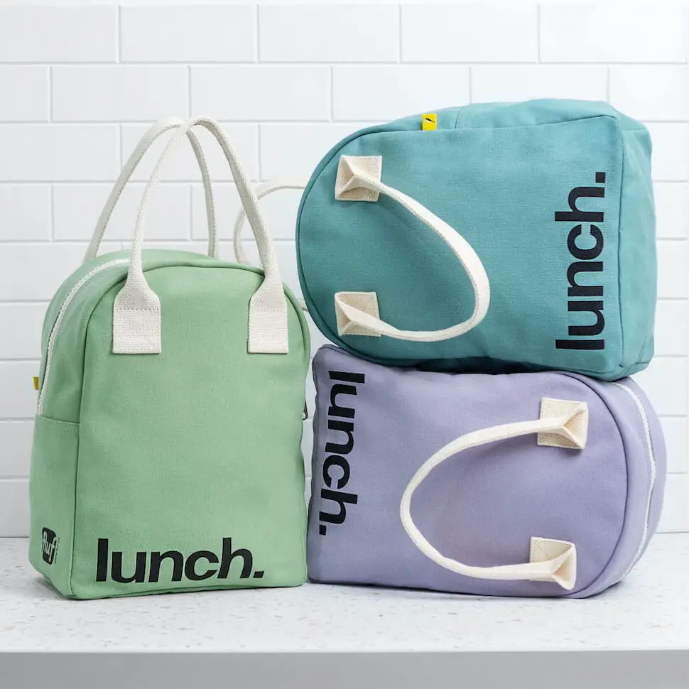 Three soft sided lunch bags with canvas handles. “lunch” is printed on the bottom left corner in black. Green, lavender, and teal bags on white brick background.