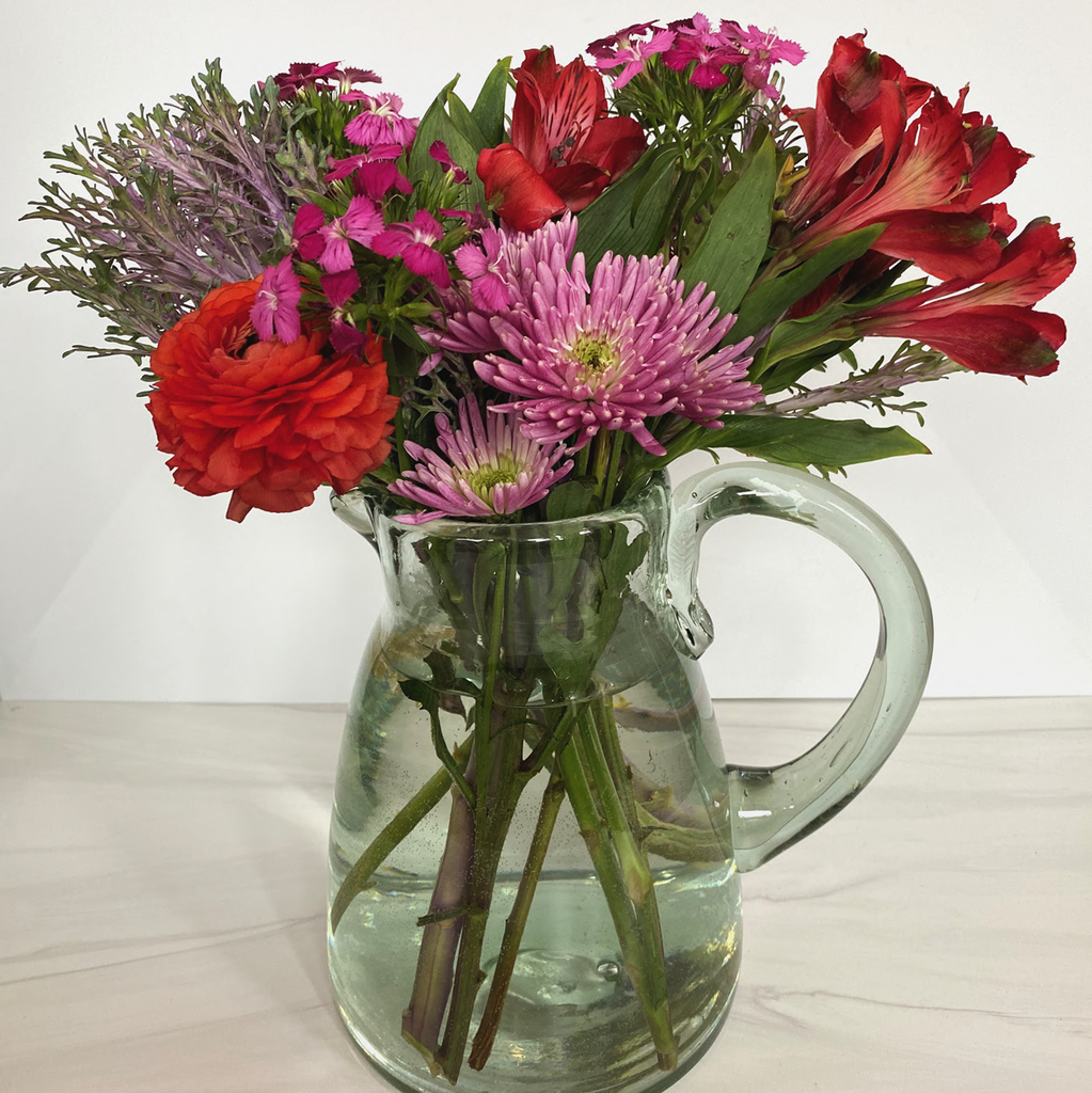 Handblown glass pitcher holding a bouquet of warm colored flowers. Classically shaped. White background.